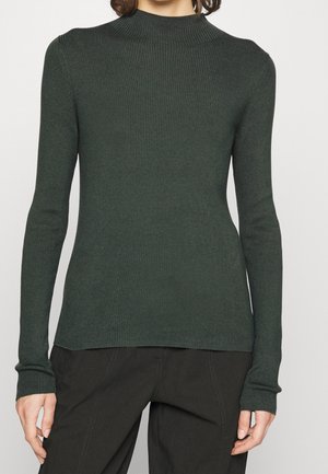 Person trägt einen dunkelgrünen, gerippten Rollkragenpullover mit langen Ärmeln und schwarze Hose, steht vor einem schlichten hellen Hintergrund.