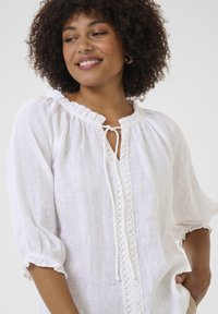 Witte linnen blouse met een rimpelende halslijn, een sleutelgatopening en decoratieve stiksels aan de voorkant. Bollende mouwen met elastische manchetten.