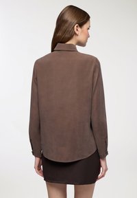Camicia marrone a maniche lunghe con una texture morbida, polsini abbottonati e orlo arrotondato, indossata sopra una gonna marrone scuro. Design minimalista, senza motivi.