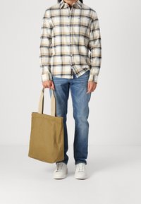 Károvaná košile v béžové a navy barvě, modré džíny a khaki plátěná taška se světlými popruhy, stojící na neutrálním pozadí.