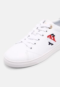 White lace-up sneaker made of smooth synthetic material. Features embroidered red, black, and white logo on the side and gold hardware accents.