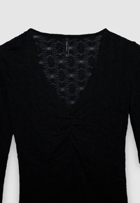 Haut noir à manches longues avec un décolleté en V, un tissu texturé à motifs floraux et un détail froncé au centre de la poitrine.