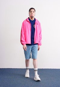 Pink athletic jacket with black stripes, blue t-shirt, denim shorts, and white sneakers with black-striped socks, standing on blue carpet.