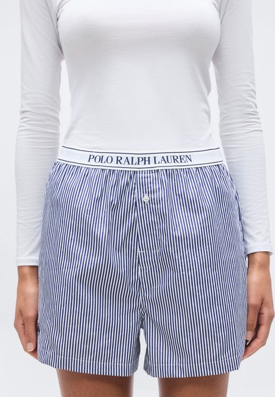 Femme portant un short à rayures verticales bleues et blanches avec une ceinture blanche portant l'inscription "Polo Ralph Lauren" et un haut ajusté à manches longues blanc.