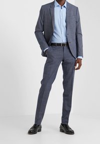 Blauw geruit pak met een lichtblauw overhemd. Het jasje heeft notch-lapels en een zak. Smalle broek met een zwart gevlochten riem en zwarte schoenen.