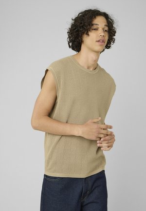 Jeune homme aux cheveux bouclés portant un haut en maille beige sans manches et un jean bleu foncé sur un fond gris uni.