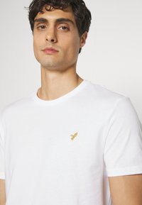 Biela bavlnená tričko s okrúhlym výstrihom a krátkymi rukávmi, s malým vyšitým zlatým logom vtáka na ľavej strane hrudníka.