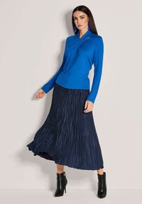 Blaues langärmeliges Oberteil mit drapiertem Ausschnitt, kombiniert mit einem gefältelten marineblauen Rock und schwarzen Knöchelstiefeln. Der Rocksaum reicht leicht unter die Waden.