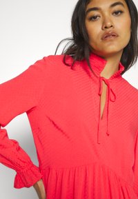 Φωτεινό κοραλλί φόρεμα με υφασμάτινη επιφάνεια, που διαθέτει ψηλό λαιμό, κλείσιμο με κορδόνι, μακριά μανίκια με πτυχές στους καρπούς, και φούστα σε φαρδιά γραμμή.