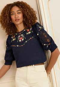 Navy blue cropped top with floral embroidery and lace accents. Short puff sleeves, ribbed cuffs, and textured fabric. Styled with cream pants.