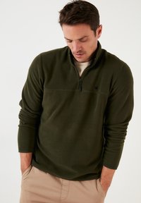 Pullover in pile verde scuro con collare a mezza zip, caratterizzato da una cucitura sul petto discreta e un piccolo logo a sinistra, vestibilità rilassata.