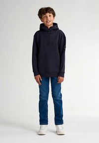 Sudadera con capucha azul marino con cierre de medio cremallera, bolsillo delantero y parche con logo en la manga, combinada con jeans azules y zapatillas blancas.