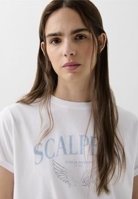 Witte katoenen t-shirt met blauwe tekst "SCALPER" en zilveren vleugelgraphics. Korte mouwen en ronde hals. Zachte textuur. Effen achtergrond.