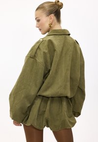 Femme aux cheveux blonds attachés en chignon, portant une veste oversize vert olive et un short assorti, vue de profil sur fond blanc.