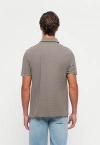 Mann iført en kortermet grå polo skjorte og lyseblå jeans, står med ryggen til mot en ensfarget hvit bakgrunn.
