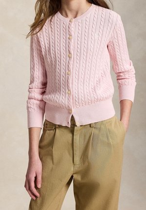 Femme portant un cardigan rose clair en maille torsadée boutonné et un pantalon beige plissé taille haute, une main dans la poche.