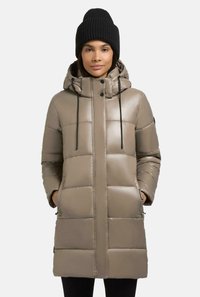 Parka imbottita di un beige chiaro con esterno in nylon, dotata di cappuccio, zip frontale, coulisse e tasche laterali. Indossata con un berretto di maglia nero.