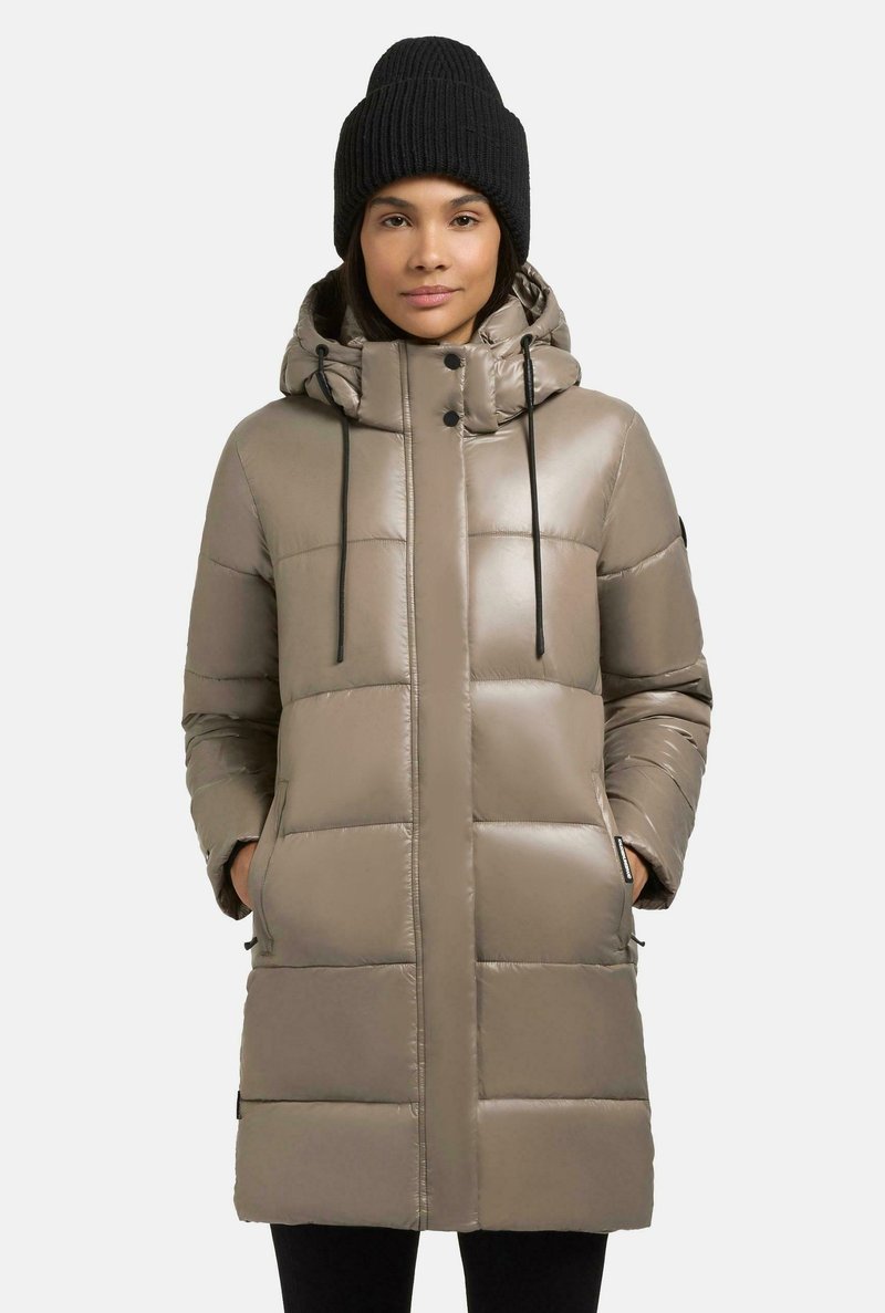 Parka imbottita di un beige chiaro con esterno in nylon, dotata di cappuccio, zip frontale, coulisse e tasche laterali. Indossata con un berretto di maglia nero.