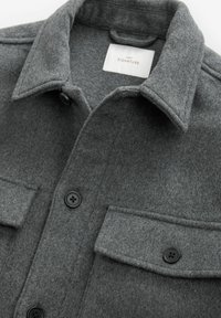 Grå skjortejakke i følget stof med spids krave, to brystlommer og sorte knaplukninger for et tekstureret og lagdelt look.