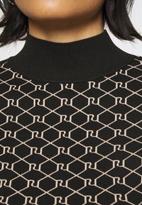 Pletený svetr s vysokým černým žebrovaným límcem a geometrickým vzorem v béžové barvě na černém pozadí. Hladká textura a přiléhavý střih.