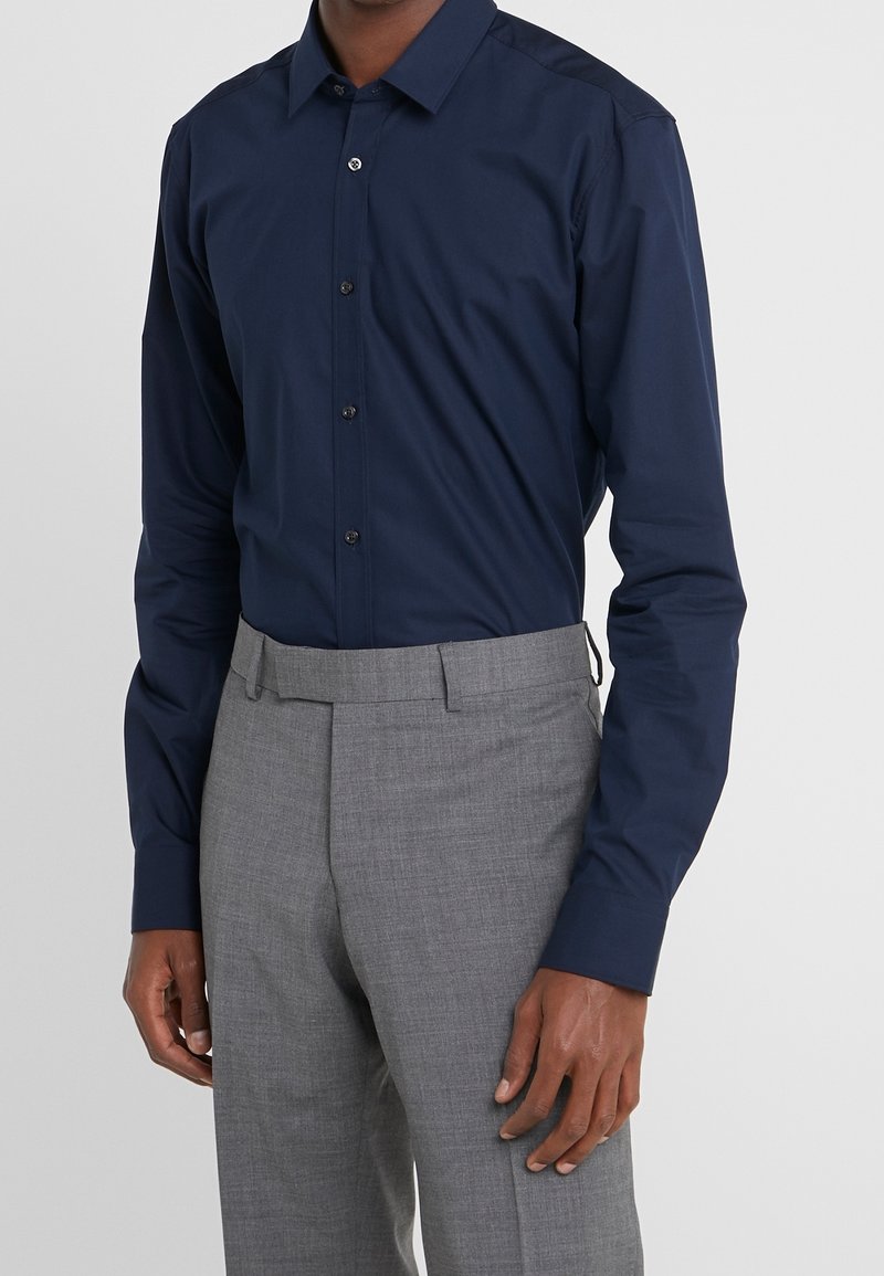 Homme portant une chemise bleu marine ajustée à boutons, rentrée dans un pantalon de costume gris clair sur mesure.