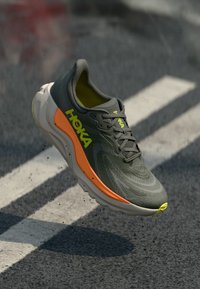 Dunkelgrüner und orangefarbener Hoka-Laufschuh in der Luft über Asphalt mit weißen Straßenmarkierungen, die Seiten- und Oberdetails zeigen.