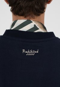 Homme portant un pull bleu marine avec le texte brodé "Interdit" dans le dos, et un col à motifs visible au-dessus de l'encolure.