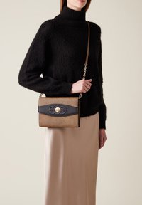 Femme portant un pull col roulé noir en peluche et une jupe beige, tenant un sac bandoulière texturé marron et noir avec des accessoires dorés.
