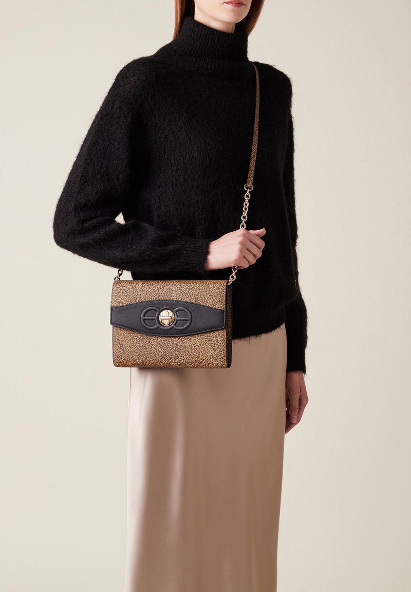 Femme portant un pull col roulé noir en peluche et une jupe beige, tenant un sac bandoulière texturé marron et noir avec des accessoires dorés.