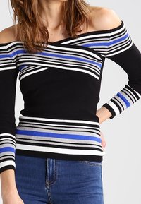 Frau in einem schwarzem, schulterfreien Pullover mit weißen, blauen und grauen horizontalen Streifen, kombiniert mit blauen Jeans.