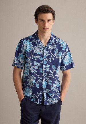Jonge man die een blauw overhemd met korte mouwen draagt met witte en lichtblauwe bloemenpatronen, staand met zijn handen in de zakken van een marineblauwe broek.