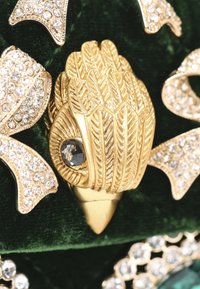 Goudkleurige broche met bladerenpatroon, voorzien van een donkere steentjesaccent. Tegen een gestructureerde groene fluwelen achtergrond.