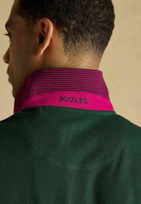 Roheline polo särk, millel on fuksia ja lilla triibuline krae. Kraele on halli värviga tikitud tekst "JOULES". Tekstuuriga kangas, juhuslik disain.