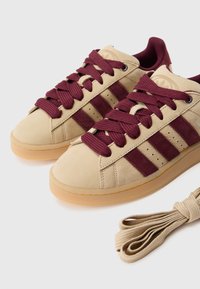 Bēša zamšādas sporta apavi ar tumšsarkaniem svītrām, apaļām auklām un gumijas zoli. Iekļautas papildu bēša krāsas auklas. Minimālistisks dizains.