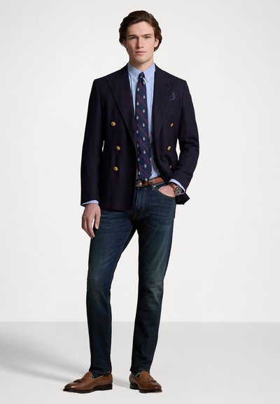Blazer doppiopetto blu navy con bottoni dorati, camicia a righe, cravatta a fantasia, jeans scuri e mocassini marroni; orologio al polso sinistro.