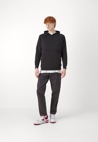 Felpa nera con cappuccio e pannello frontale, abbinata a pantaloni cargo grigi con tasche laterali e sneakers bianche con dettagli rossi.