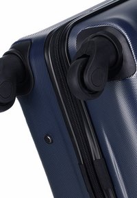 Hard-shell suitcase in dark blue with a textured finish, featuring black wheels and zipper, and a small black button detail beside the zipper.