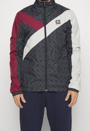 Homme portant une veste à zip noire avec des motifs circulaires répétitifs, une manche gauche bordeaux, une bande diagonale blanche et le logo ellesse sur la poitrine.