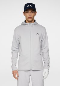 Grauer Reißverschluss-Sweatshirt mit Kapuze und schwarzem Logo, kombiniert mit hellgrauen Hosen und einer marineblauen Mütze. Hat eine glatte Textur und seitliche Taschen.