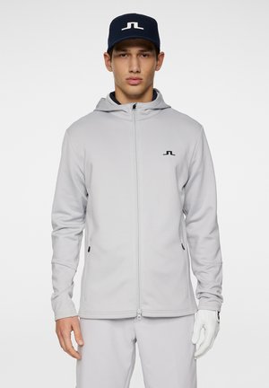 Felpa grigia con zip e cappuccio, abbinata a pantaloni grigi chiari e berretto blu navy. Presenta una texture morbida e tasche laterali.