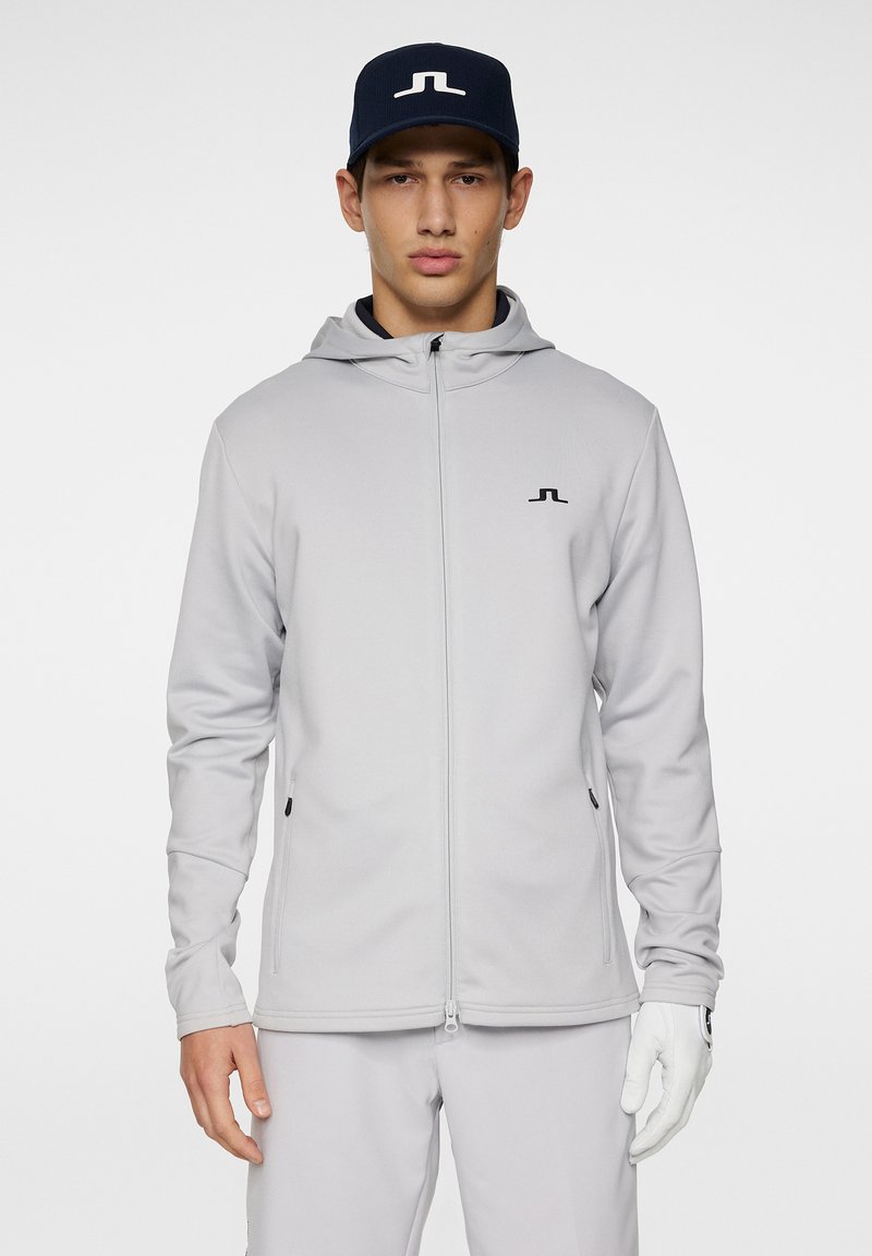 Grauer Reißverschluss-Sweatshirt mit Kapuze und schwarzem Logo, kombiniert mit hellgrauen Hosen und einer marineblauen Mütze. Hat eine glatte Textur und seitliche Taschen.