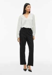 Weiße Bluse mit langen Ärmeln und einem tiefen V-Ausschnitt, kombiniert mit hochtaillierten schwarzen Hosen und schwarzen spitzen Absatzschuhen. Glatte Stoffoberfläche.