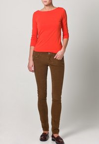 Rode top met lange mouwen en boothals, gecombineerd met bruine skinny broek van ribfluweel, voorzien van zakken aan de voorkant en een knoop. Draagt daarbij donkere loafers.