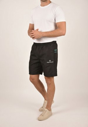 Hombre con camiseta blanca, pantalones cortos negros Sergio Tacchini con franjas laterales y zapatillas beige, de pie con las manos entrelazadas.