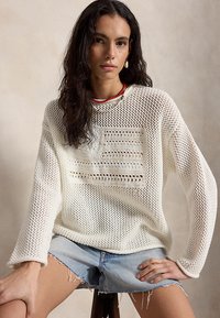 Γυναίκα με μακριά σκούρα μαλλιά φοράει λευκό πλεκτό πουλόβερ με σχέδιο σημαίας, ανοιχτόχρωμα τζιν σορτς, κόκκινο κολιέ με χάντρες και ένα σκουλαρίκι, καθιστή.