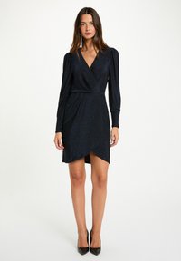 Femme debout portant une robe enveloppante sombre et scintillante avec de longues manches bouffantes et des talons aiguilles noirs, sur un fond blanc uni.