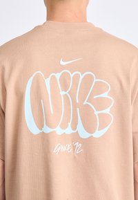 Bež pamučna majica s velikim plavim grafitnim logotipom "NIKE" i natpisom "SINCE '72", uz bijeli Nike swoosh iznad dizajna.