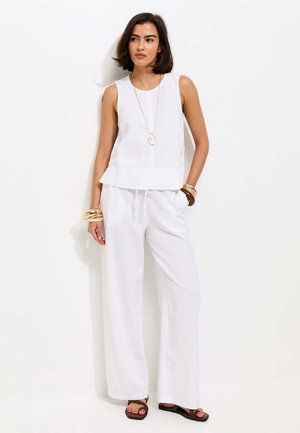 Kobieta o krótkich ciemnych włosach, ubrana w białą bluzkę bez rękawów, szerokie białe spodnie, sandały, długi naszyjnik oraz kilka bransoletek, stoi przy białej ścianie.