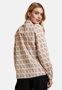 Langärmlige Bluse aus einem cremefarbenen Stoff mit einem Spielkartenmuster in Schwarz und Rot. Gerader Schnitt mit Knopfdetails an den Ärmeln.