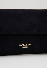 Pochette en daim noir avec logo "Dune London" doré centré sur le rabat avant sur un fond uni.